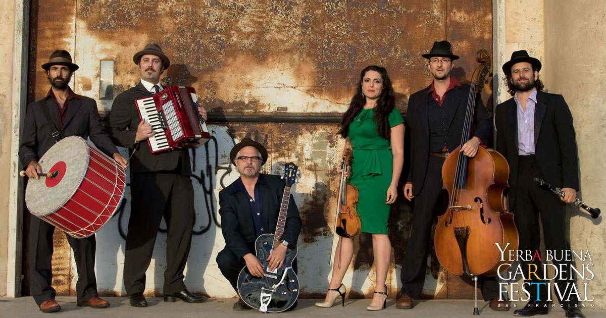 Photo of klezmer music group Kugelplex, with band members standing lined up holding their instruments in front of a worn industrial door