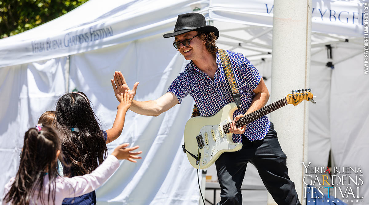 A guitarist with a hat and sunglasses, on stage, smiling and high-fiving a young audience member
