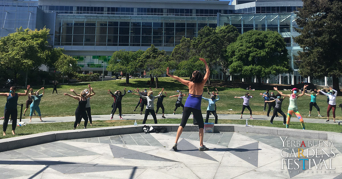Back of a dance instructor on the Yerba Buena Gardens Festival Stone Stage, arms stretched upwards. Facing a group of dance class students standing on the grass lawn, also with their arms stretched out.
