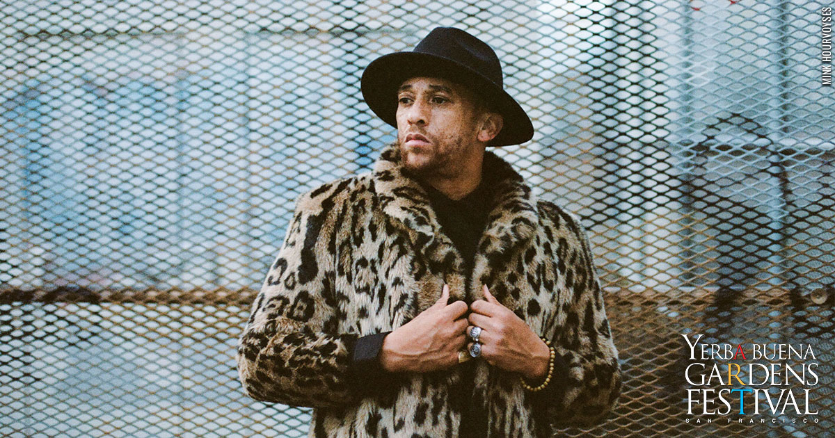 Kev Choice outside in an industrial warehouse area, against a large fence. Wearing a black fedora hat and leopard print fur coat.