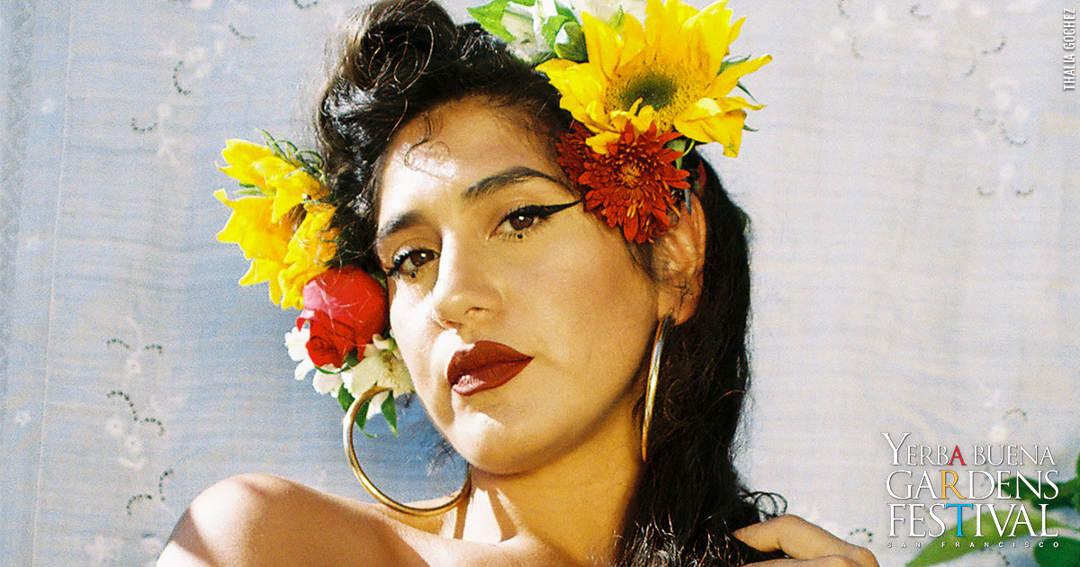Headshot photo of La Doña in a white top, wearing big gold hoop earrings and red and yellow flowers in her hair.