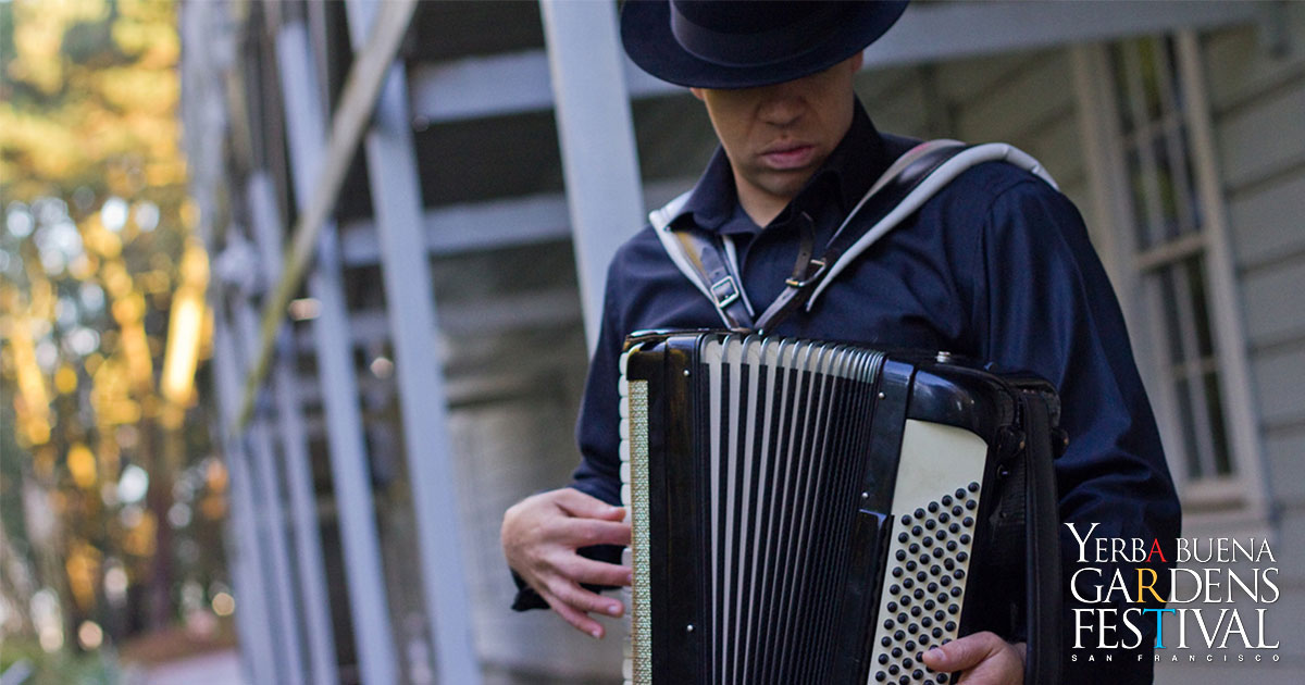 André Thierry looking down, to the right, face covered by a black fedora hat as he holds and plays and accordion