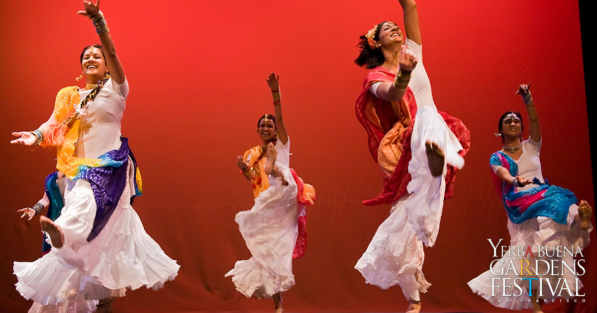 Four Dholrhythms Dance Company dancers, staggered on a stage with a red backdrop, in the middle of a hopping motion.