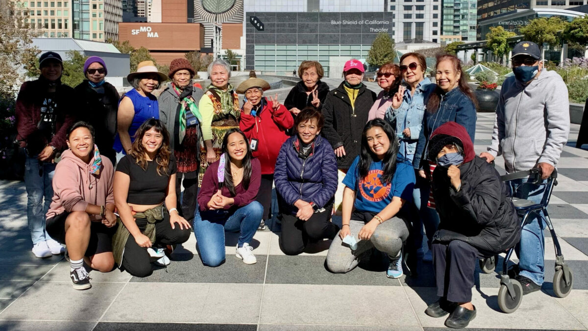 Members of Bindlestiff Studio's Restorative Theater Arts for Seniors program gathered together, smiling and posing on the Yerba Buena Gardens Terrace in the bright sunshine.