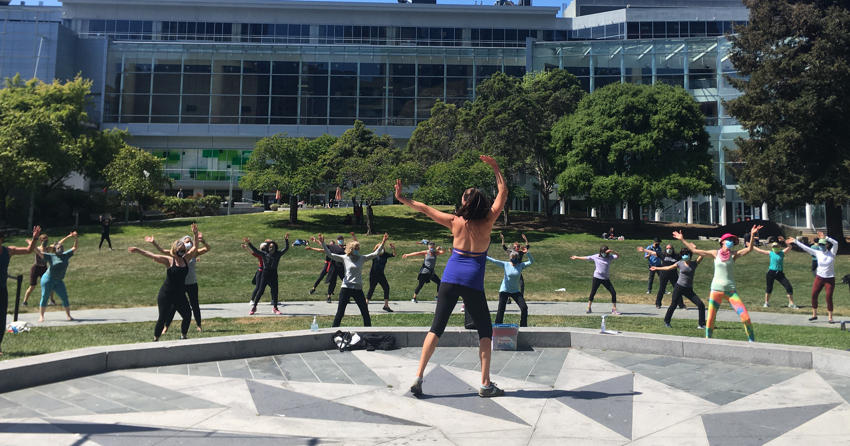 Back of a dance instructor on the Yerba Buena Gardens Festival Stone Stage, arms stretched upwards. Facing a group of dance class students standing on the grass lawn, also with their arms stretched out.