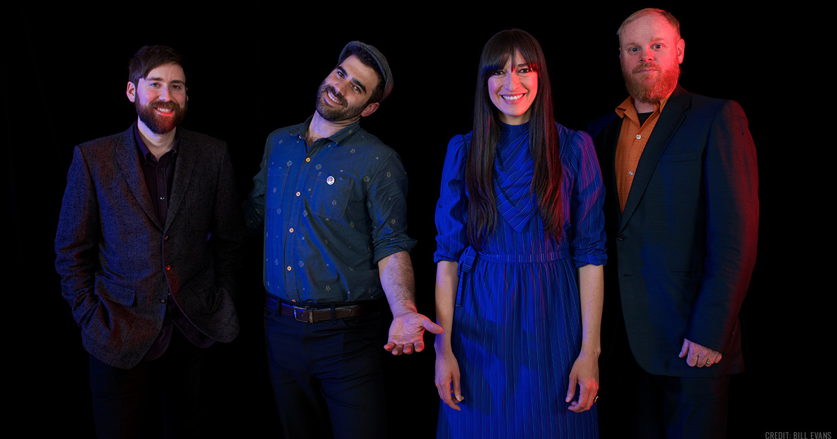 Diana Gameros with her band, Patrick Wolff, Thomas Edler and Aaron Kierbel, standing and smiling against a dark backdrop.