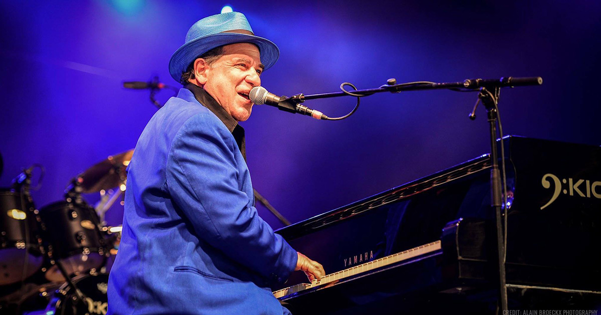 Mitch Woods in blue suit and fedora, sing and playing piano on a stage in front of his Rocket 88's band.