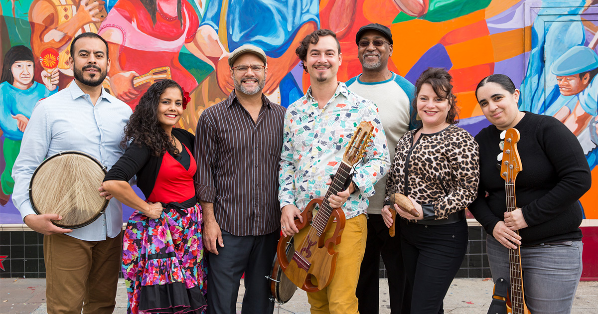 Members of La Mixta Criolla smiling and holding their instruments, in front of a colorful mural