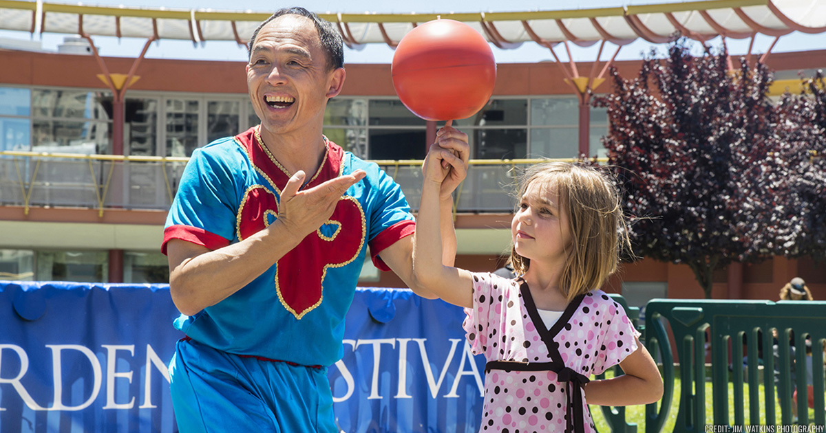 Wayne Huey of the Red Panda Acrobats smiling whiling helping a young child spin a ball on their finger.