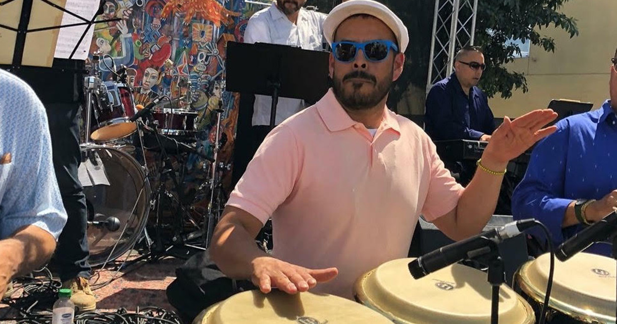 Javier Navarette wearing shades and a hat playing congo drums outdoors with a band.