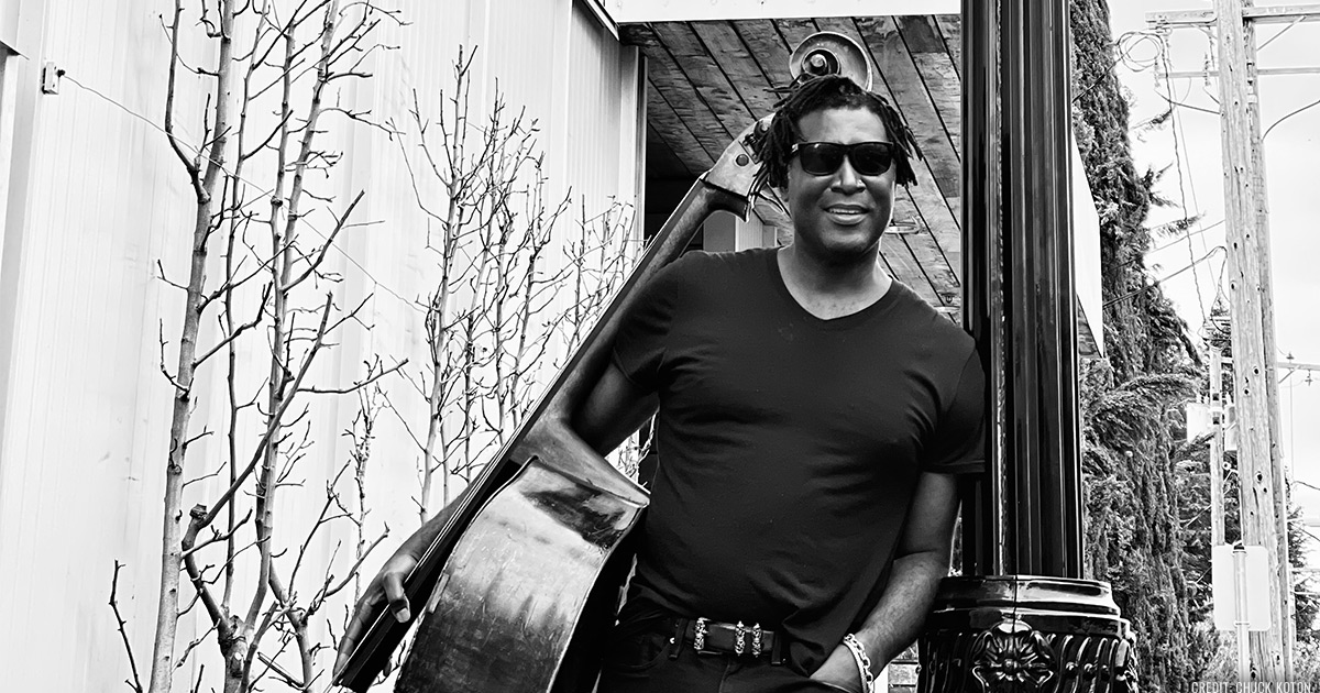 Black and white photo of Marcus Shelby leaning his left arm on a light post and holding an upright bass on under his right arm.