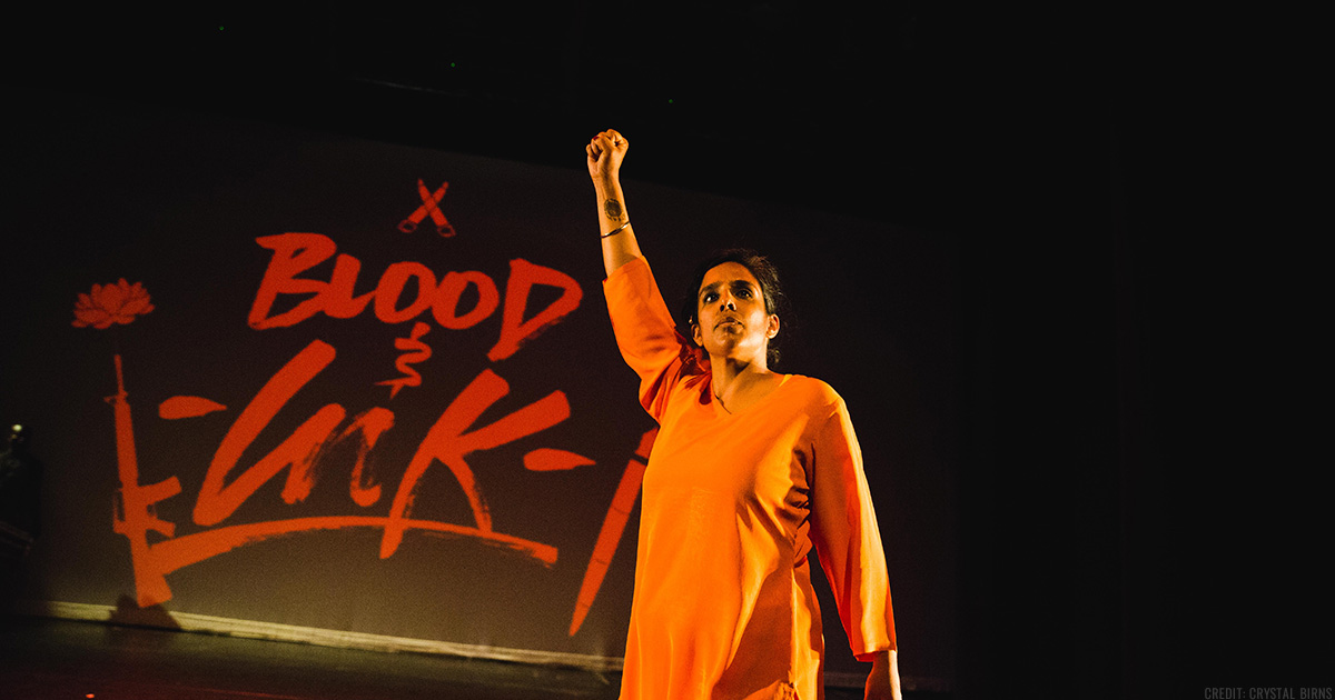 Joti Singh on a theater stage wearing an orange outfit with her right fist raised in the air, the words "Blood Ink" projected in red on the screen behind her