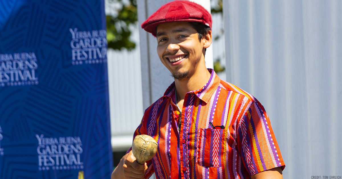 Ahkeel Mestayer wearing a brightly colored shirt and hat, smiling and holding maracas in his hand on the YBG Festival stage, a blue banner behind him.