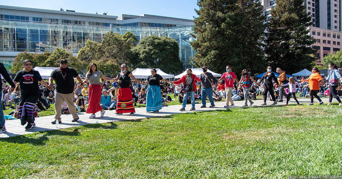 Performers and audiences from Indigenous People's day part of a friendship dance, holding hands and walking through the Yerba Buena Gardens lawn