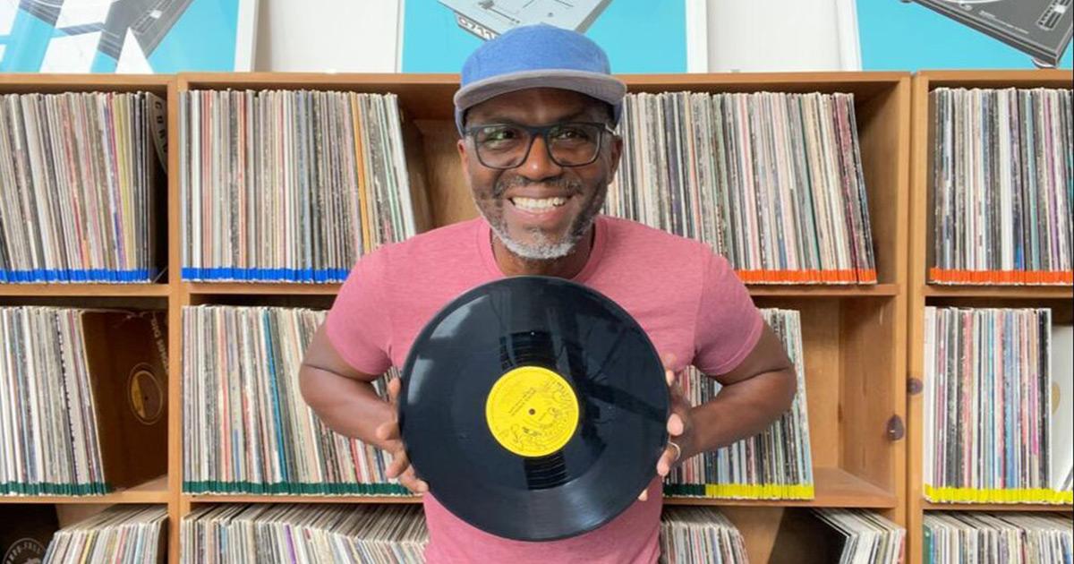 DJ Lamont smiling and holding a vinyl record, standing in front of shelves packed with vinyl covers