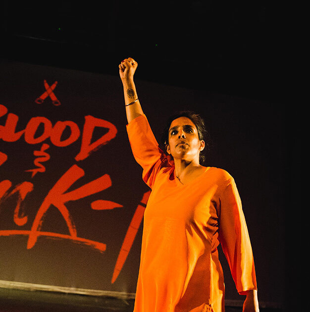 Joti Singh on a theater stage wearing an orange outfit with her right fist raised in the air, the words "Blood Ink" projected in red on the screen behind her