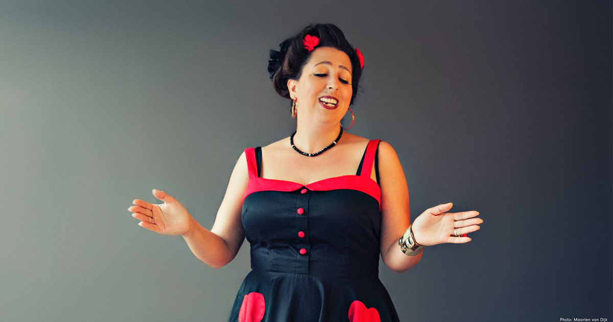 Jewlia Eisenberg in a black and red dress, singing in front of a grey backdrop