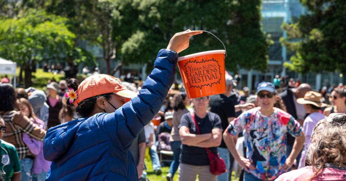 YBG Festival staff member holding up orange donation bucket for audiences at a an outdoor event.