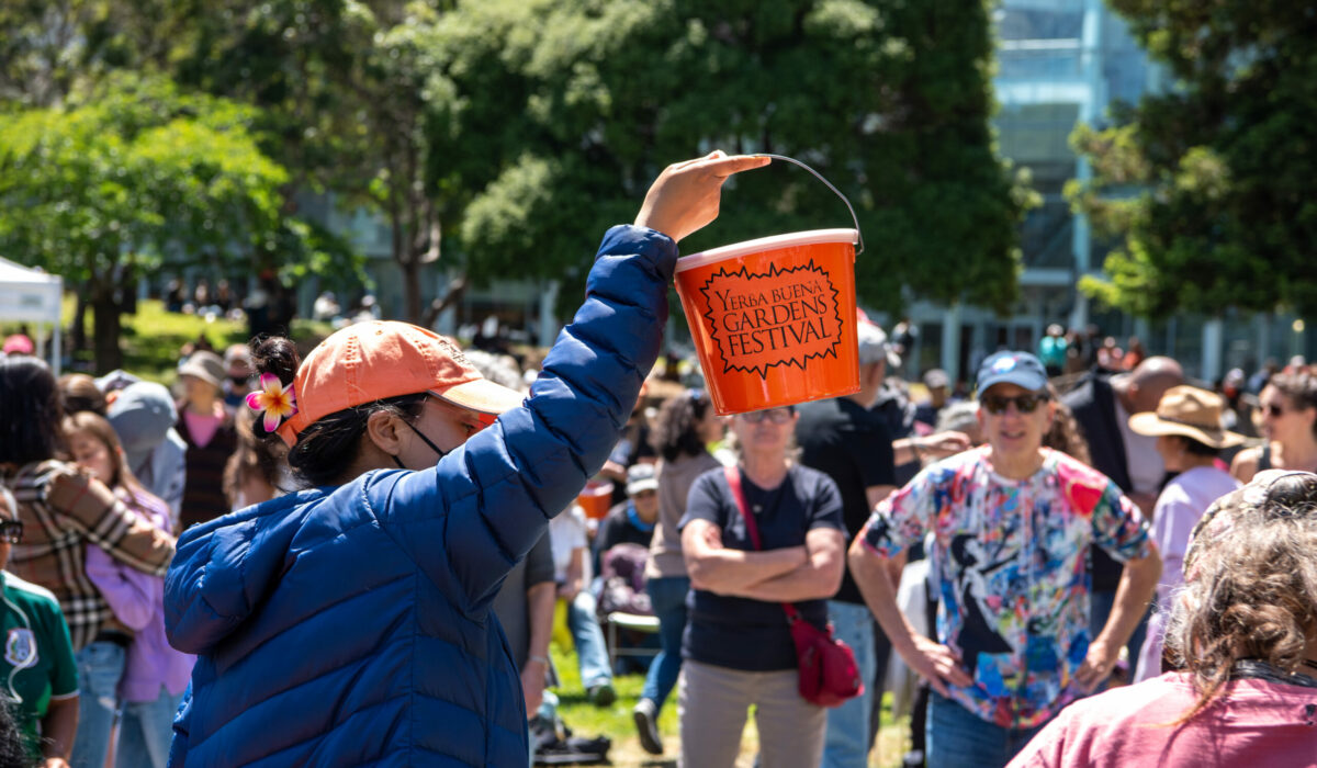 YBG Festival staff member holding up orange donation bucket for audiences at a an outdoor event.