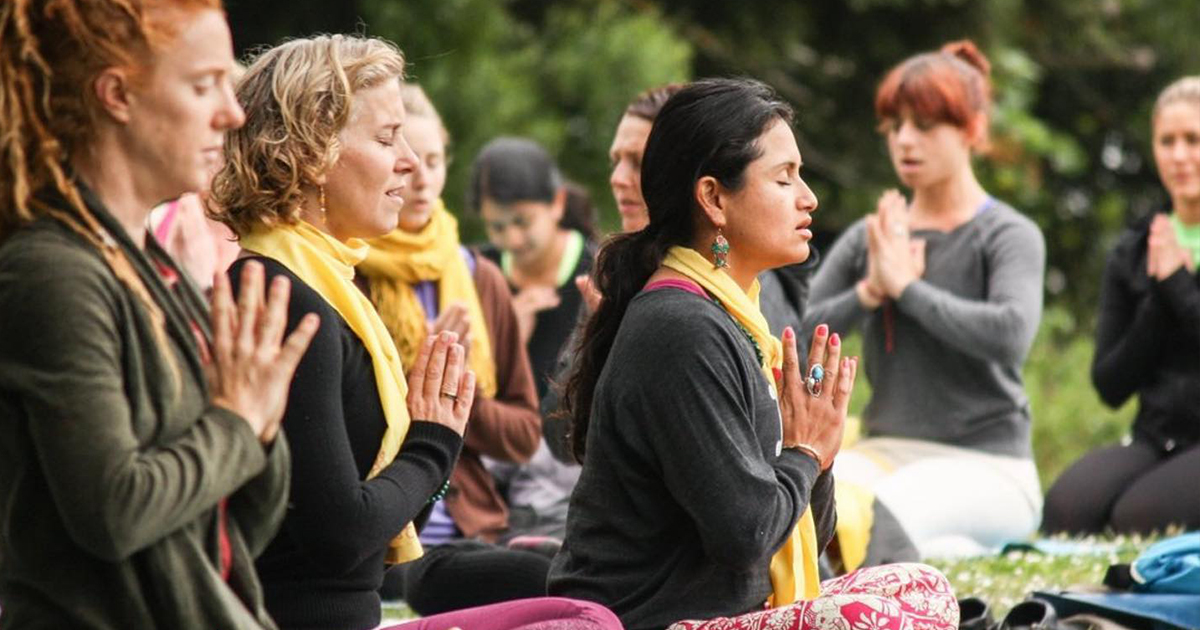 A group of people sitting outside, their eyes closed and hands in prayer position for yoga meditation.