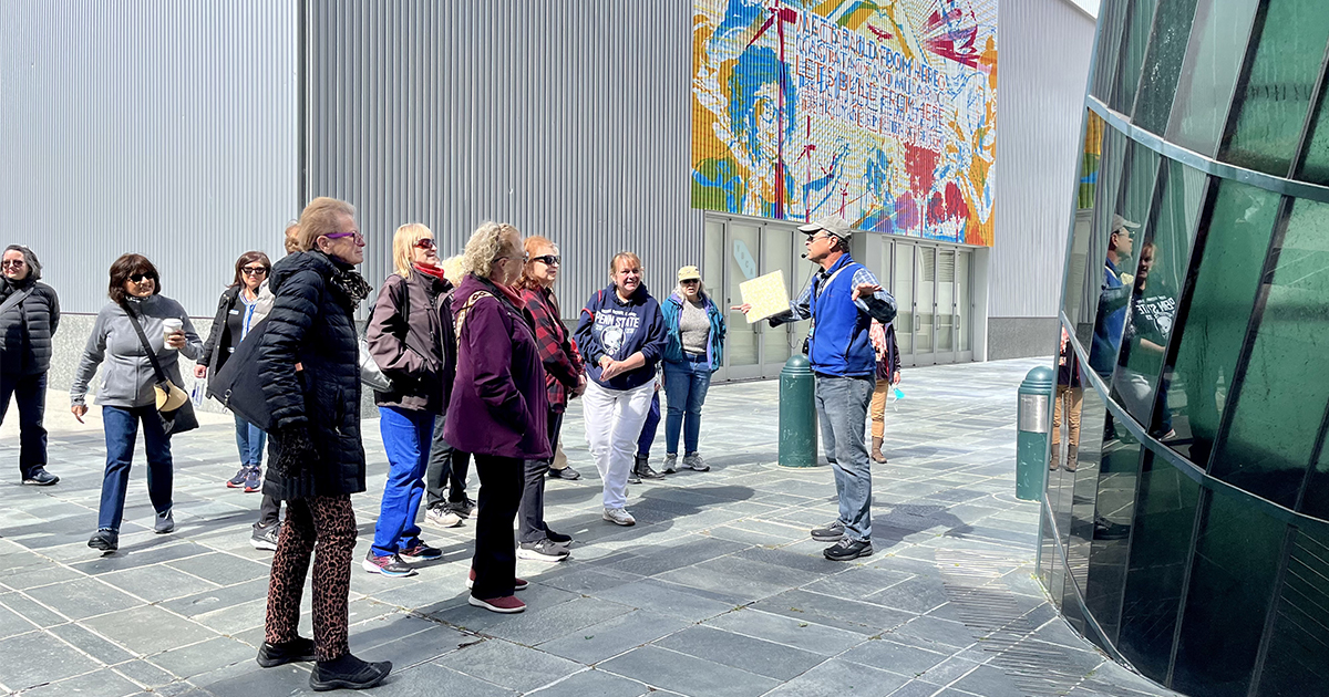 A group of people gathering with a tour guide outside in front of a large public art statue, behind them a colorful mural on the grey wall.
