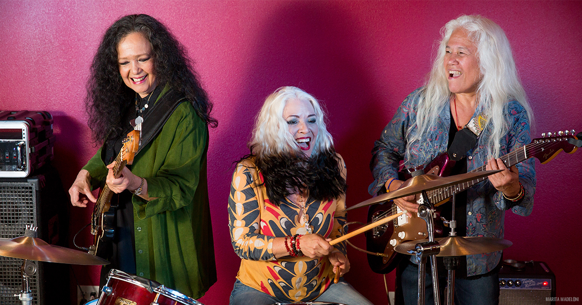 3 members of Fanny all smiling and playing instruments in front of a magenta backdrop.