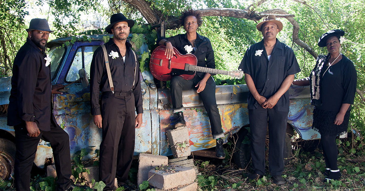 Anita Lofton holding a guitar, members of her band on both her sides, leaning on an old truck under green leafed trees