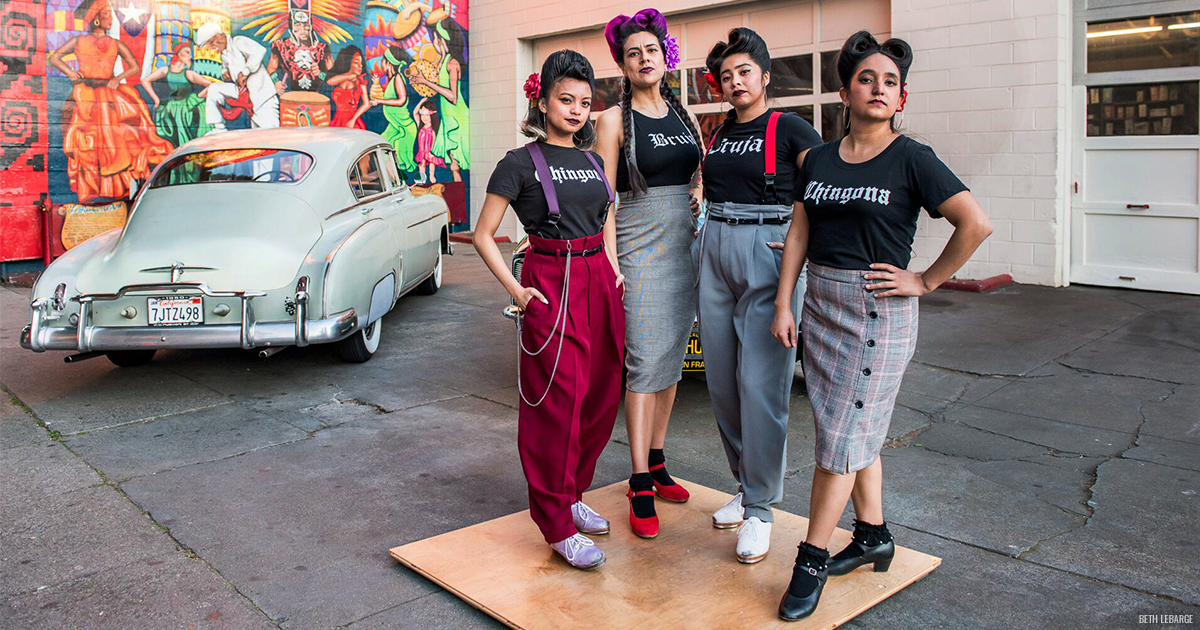 4 members of La Mezcla standing on a square tap dance board, outside in front of a vintage car.