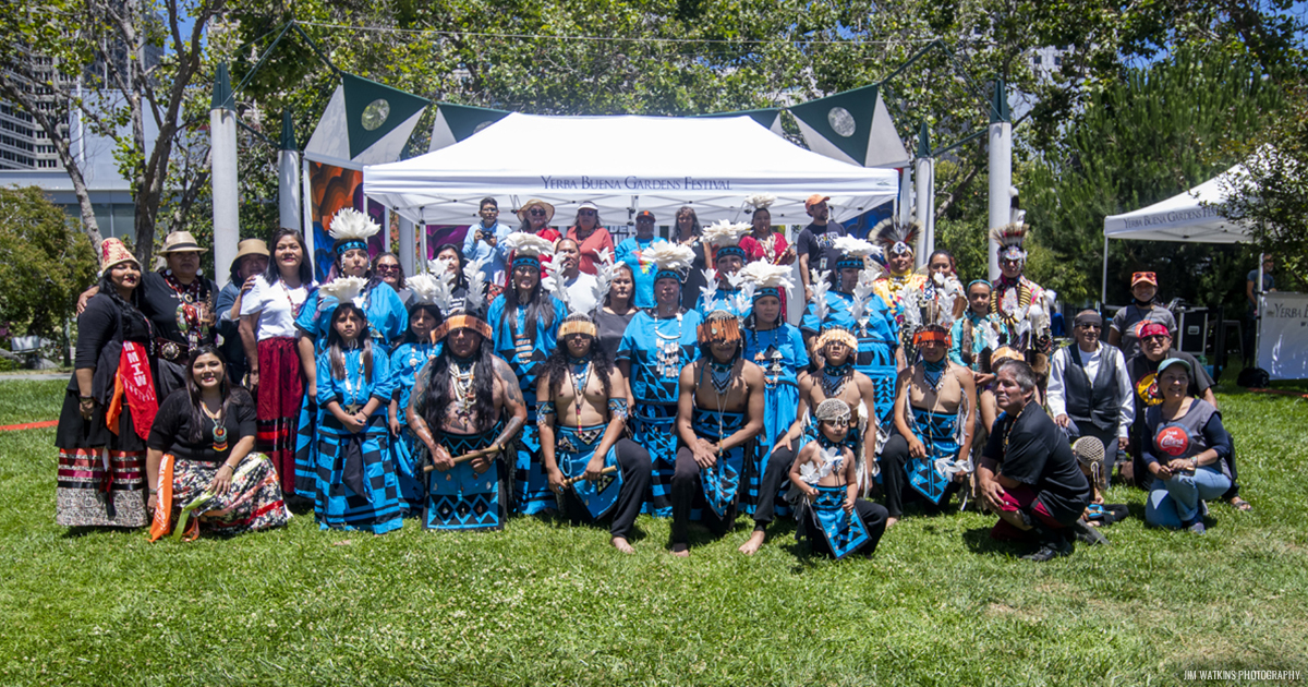 A few dozen Native artists and performers dressed in traditional regalia, standing outside on a green lawn.