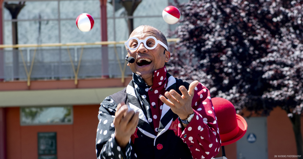 Unique Derique wearing a bright red, white and blue outfit, juggling two balls in the air.