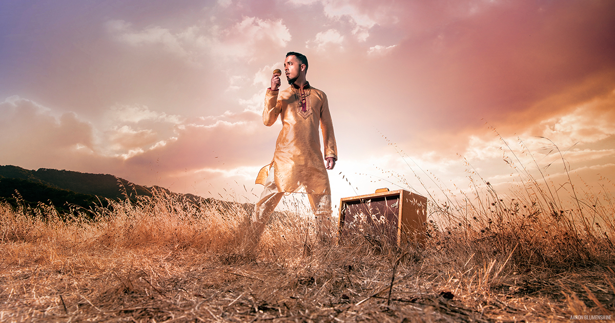 Singer Aki Kumar in a golden traditional Indian, wardrobe standing in the middle of a golden field, holding a microphone with the clouds and sun behind him