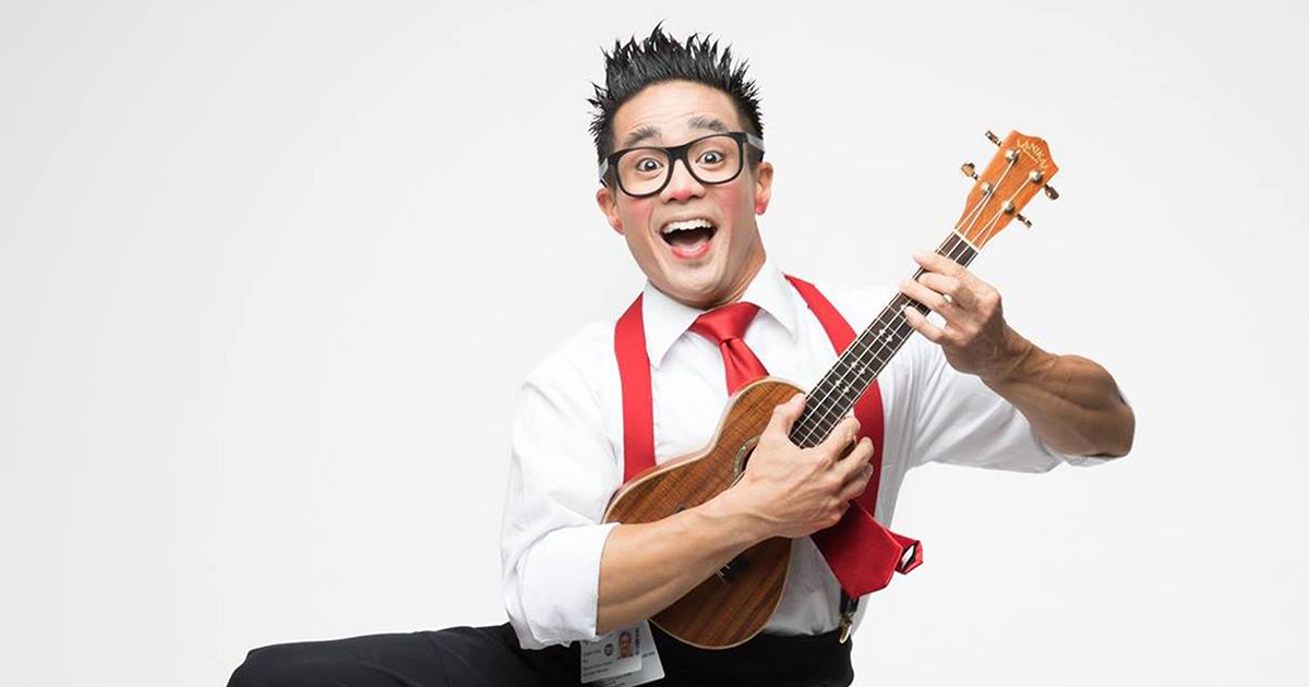 Calvin Kai Ku wearing a white shirt and red tie and suspenders, in a leaping motion, smiling and holding a ukulele