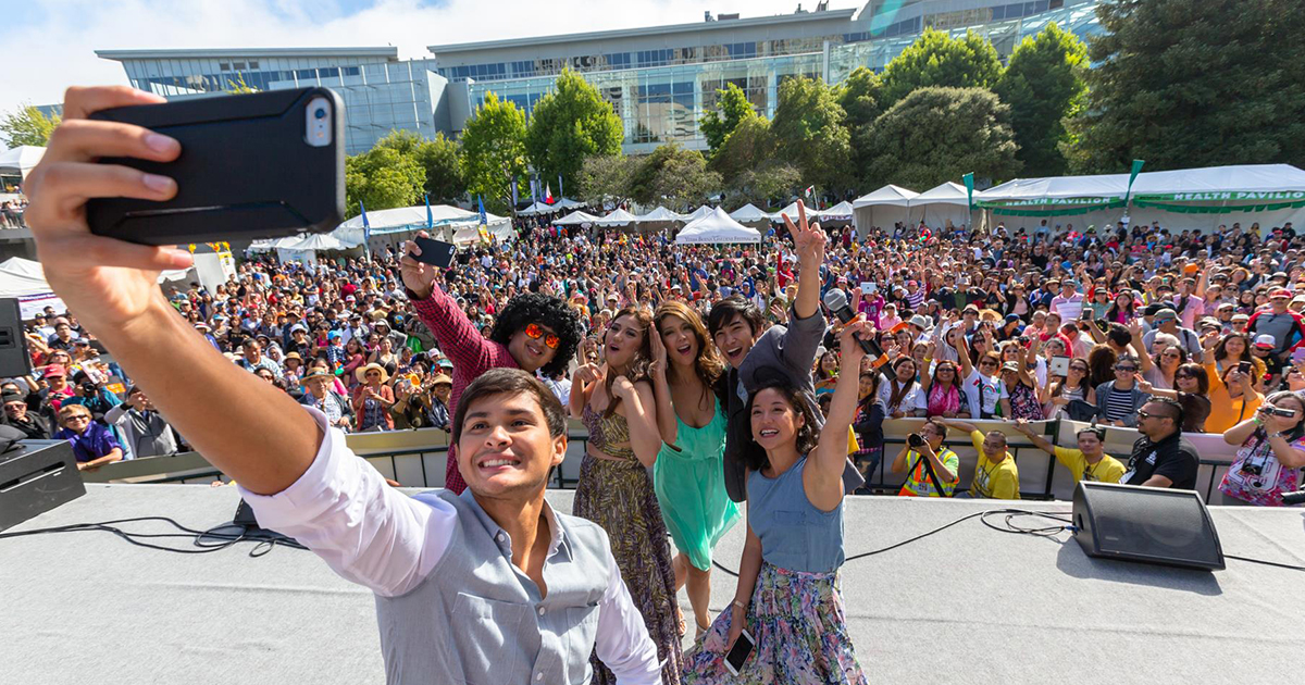 4 people on a stage taking a selfie photo with a large crowd of audiences behind them off-stage