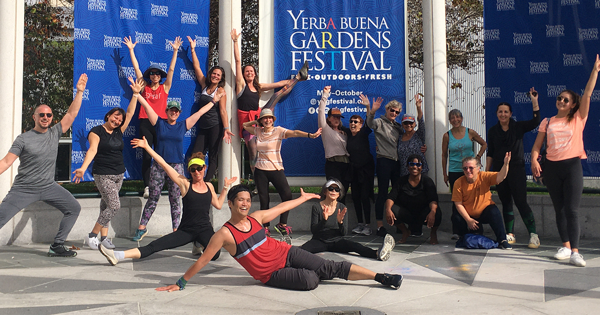 A group of the Dance Outdoors with Rhythm & Motion class smiling and posting in front of the Yerba Buena Gardens Festival stage banner.