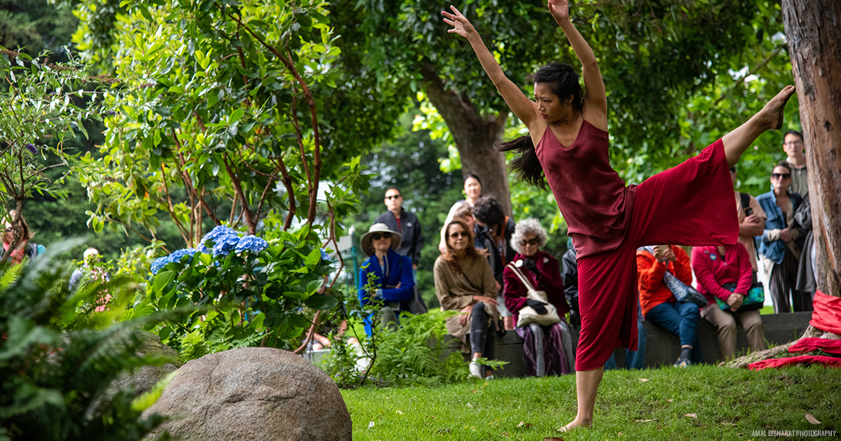 Female dance wearing red in an outstretched stance, in the middle of a lush green spot of grass and audiences sitting nearby