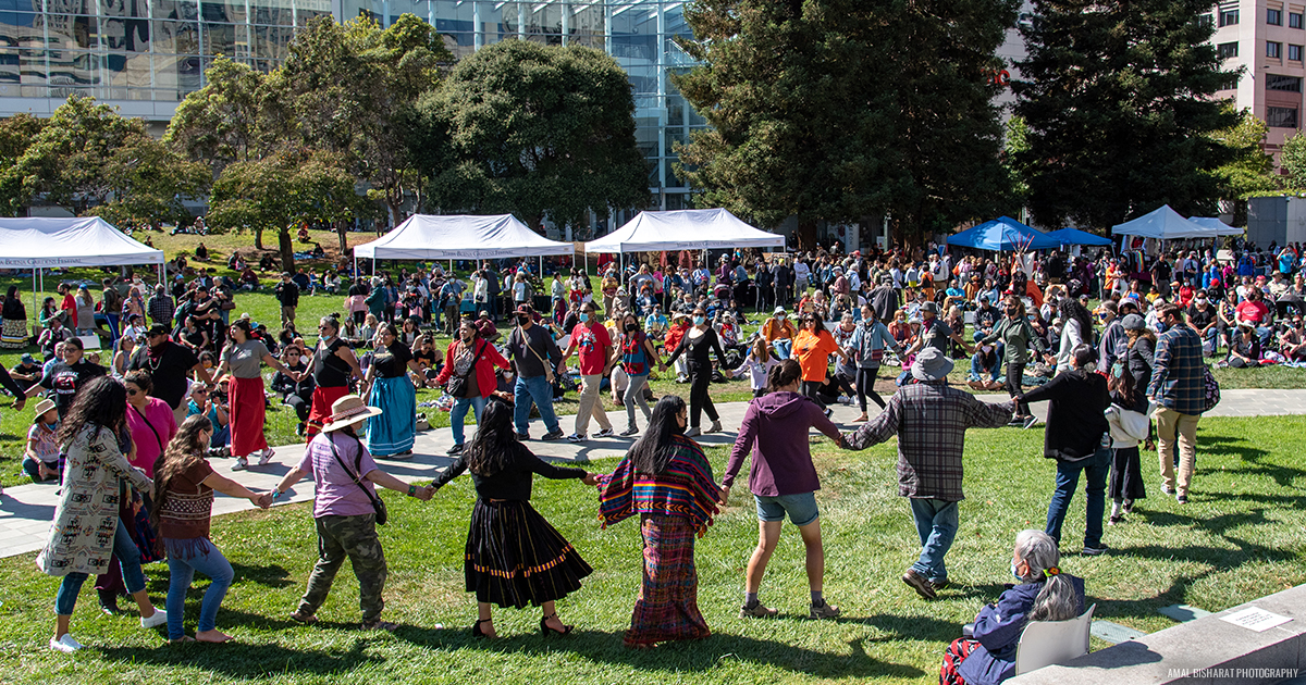 A large group of people dancing in a friendship circle in the middle of the Yerba Buena Gardens esplanade lawn.