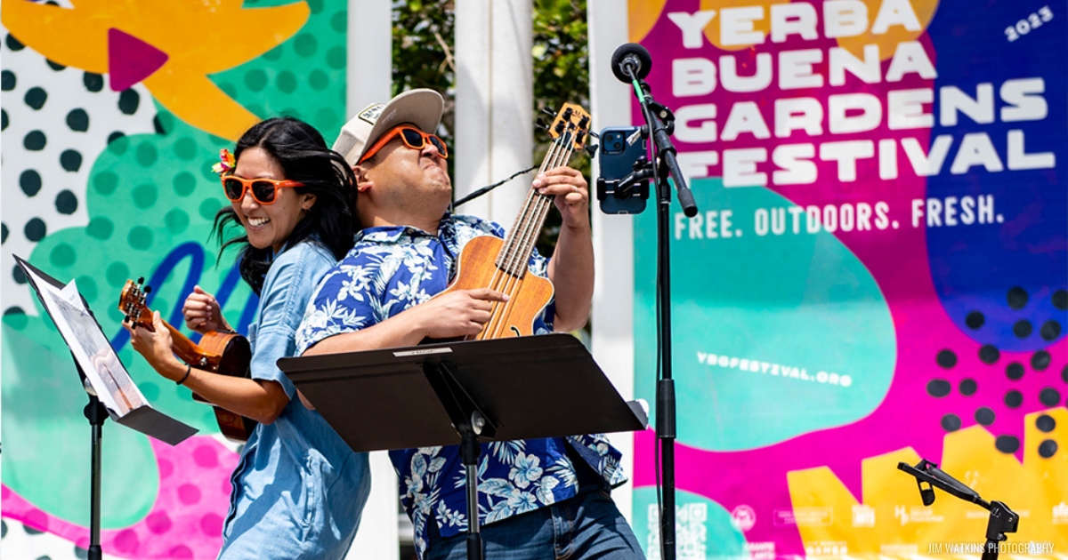 Man and woman smiling, standing back-to-back, each playing an ukulele, in front of a colorful stage backdrop