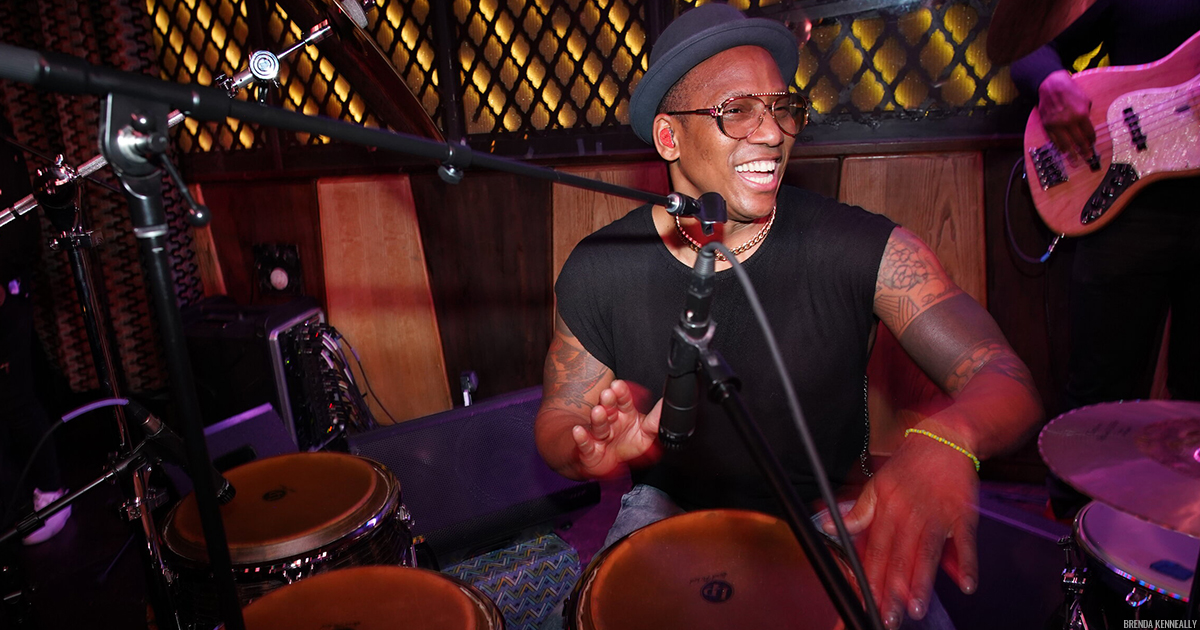 Pedrito Martinez smiling, behind microphone stands, playing a set of bata drums with his hands