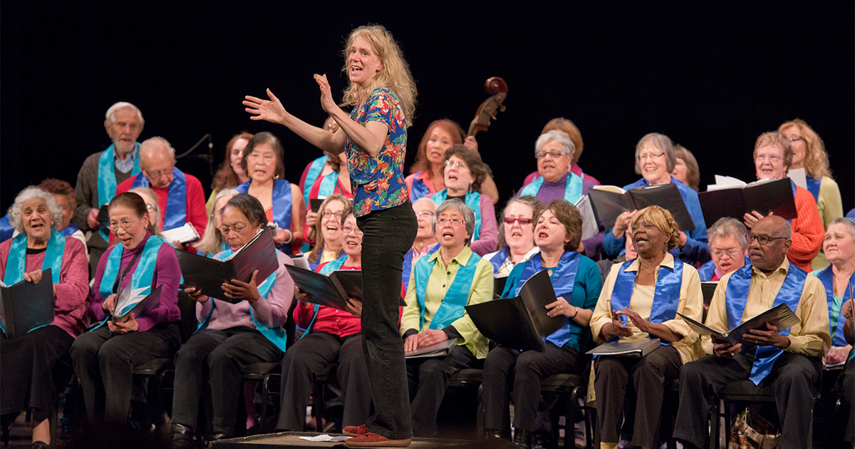 A choir group sitting and singing, each wearing a teal stole, choir director standing in front of them.