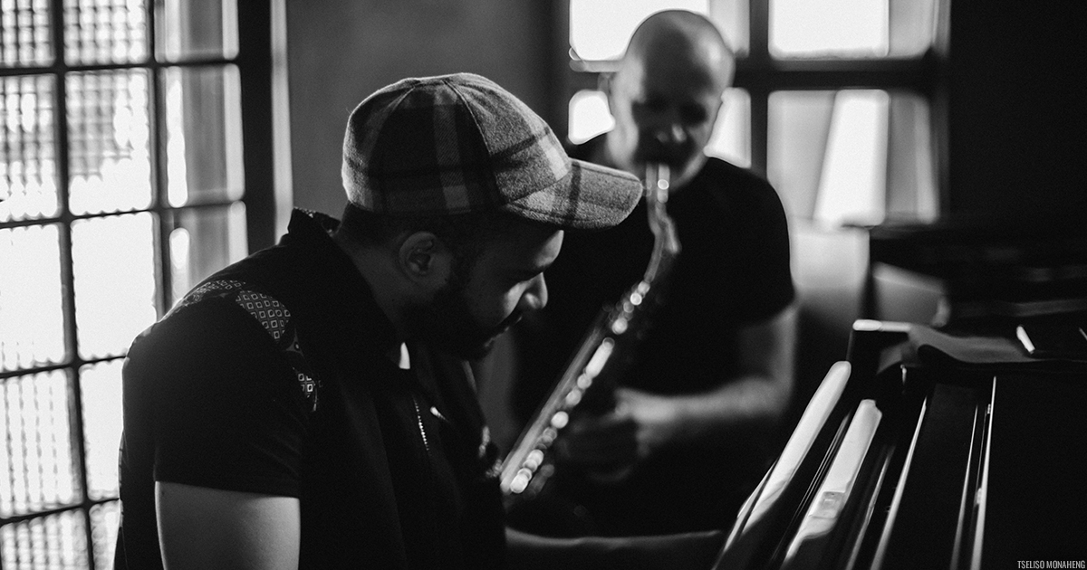 Black and white photo of a person wearing a cap, sitting at and playing the piano, behind them another person playing the saxophone.