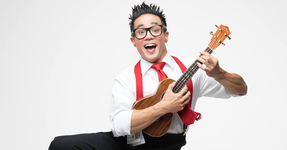 Calvin Kai Ku wearing a white shirt and red tie and suspenders, in a leaping motion, smiling and holding a ukulele