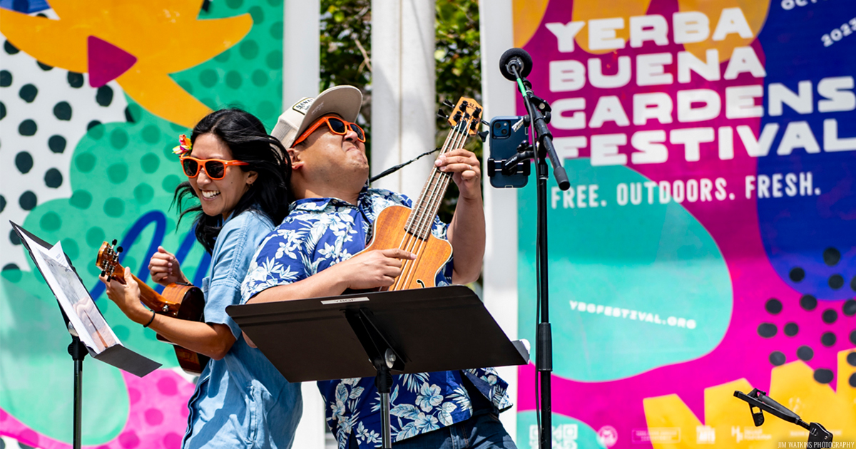 Cynthia Lin and Ukulenny leaning on each other back-to-back, wearing orange sunglasses and joyfully playing ukuleles in front of a color YBG Festival banner.