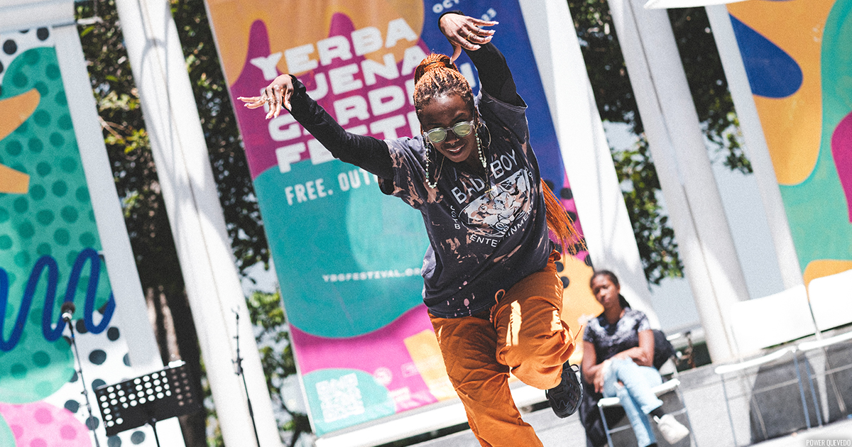 A freestyle dancer on an outdoor stage in front of a colorful YBG Festival banner.