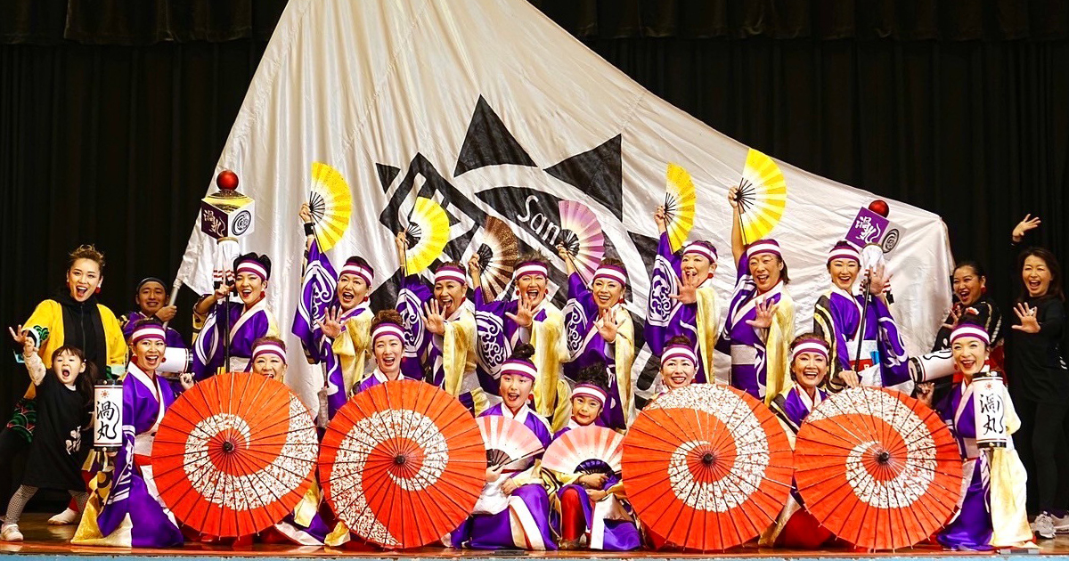 Adults and children of UZUMARU front-facing, happily yelling and smiling, wearing colorful attire, holding up parasols and fans, with a large off-white flag behind them