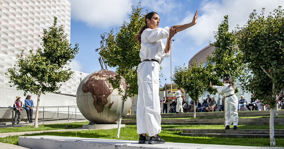 A dancer in a white jumpsuit with their arm stretched out to the right, performing on outdoors in front of a few trees and a outdoor sculpture.