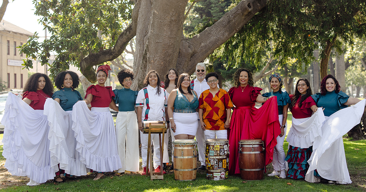 Members of Batey Tambó standing outdoors in front of a large tree, each wearing traditional Puerto Rican attire and with barriles de bomba in front of them.
