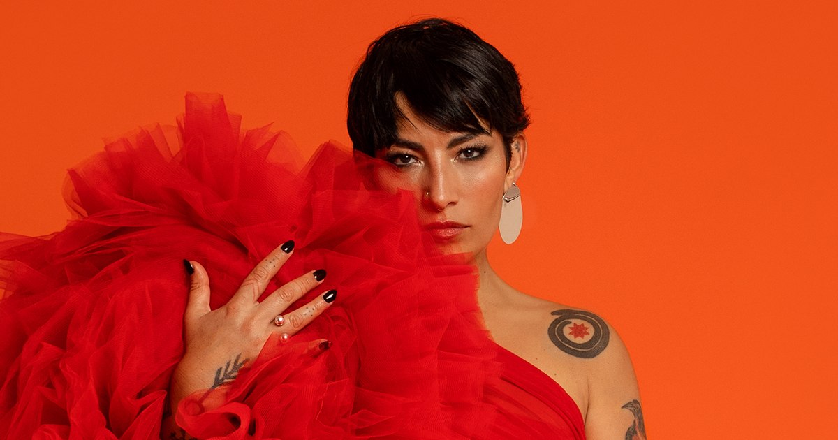 Ana Tijoux, front-facing, holding a billowing red fabric near her face with 1 hand, in front of a vibrant orange backdrop