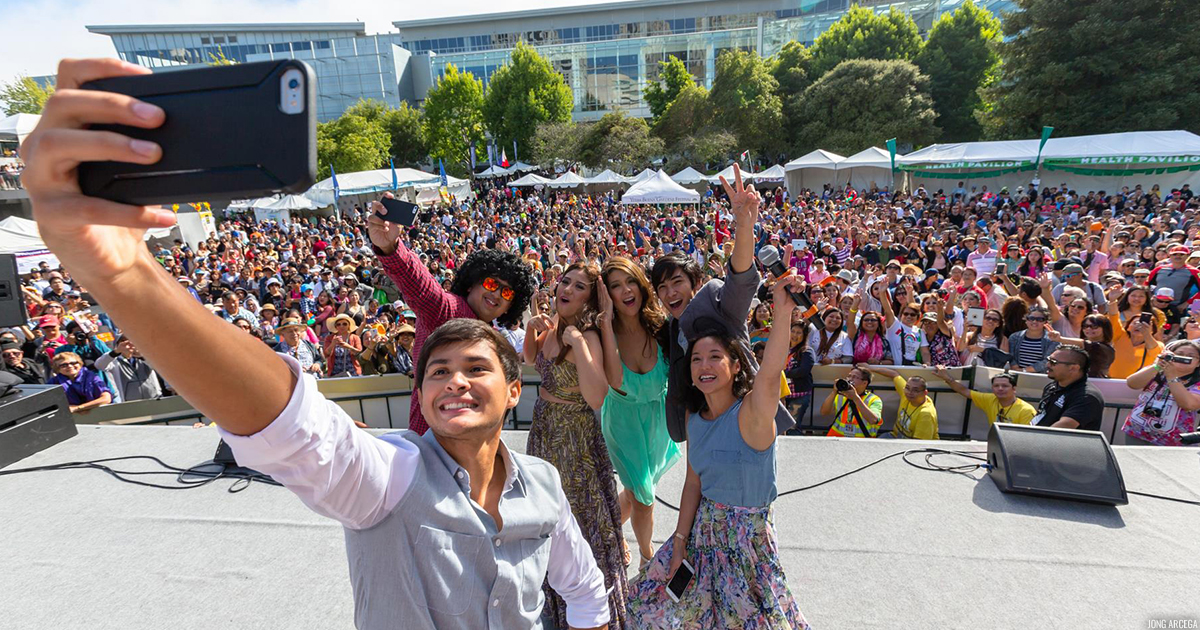 4 people on a stage taking a selfie photo with a large crowd of audiences behind them off-stage