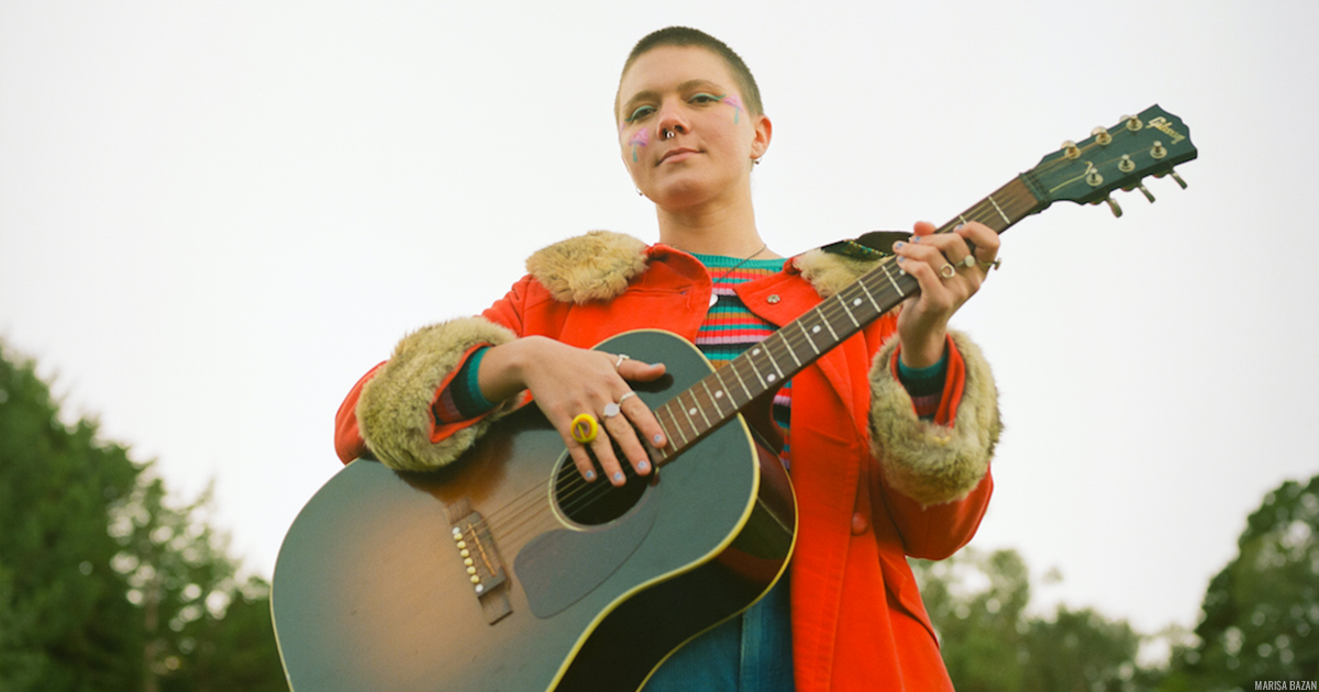 Mae Powell standing outdoors, wearing a bright orange jacket and holding a guitar in her arms.