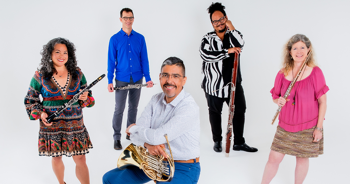 5 members of Quinteto Latino, front-facing, each smiling, some holding an instrument, standing in front of a white backdrop